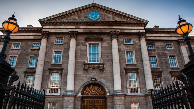 It is approaching dusk. The front of Trinity College stands tall and centre stage in the image. The old oak entrance door to Trinity College has closed for the night, above it, the blue clock with gold hands points at twenty-past seven. The old circular lamp posts in the foreground of the image are starting to light up. 
