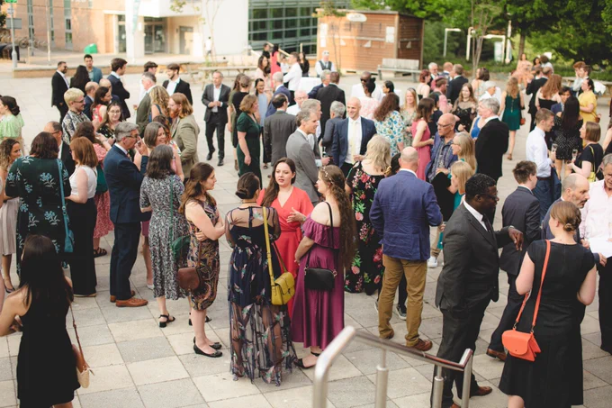 A large group of students and staff mingling outside the Forum, drinking prosecco and orange juice.