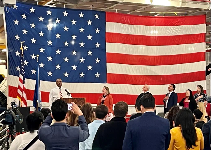 A group of people attending a naturalization ceremony face the podium at the USS Hornet Museum. A large U.S. flag hangs in the background.