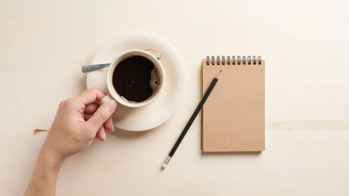 A photograph of a cup of coffee, a notebook and a pen on a white background