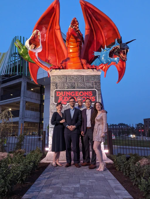 Five members of the Video Game History Foundation standing in front of a large sculpture. It depicts a section of wall with a prominent “Dungeons and Dragons” logo. On top is a dramatic  five-headed dragon sculpture that breathes fire or releases mist depending on the random action chosen.