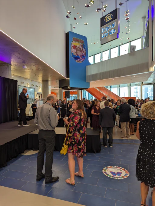 New lobby of The Strong. Steve Dubnik, CEO of The Strong stands on stage in a suit with black and white sneakers. The well-dressed crowd is milling about in front of him. The mezzanine is visible with a giant Donkey Kong arcade machine feeling like it was built into the walls and a large sign that reads ESL Digital Worlds shaped like an NES cartridge hangs from the ceiling. In the back you can see a dramatic staircase that leads up to the mezzanine level. Also visible are dozens of LED ‘butterflies’ that display information visible when using an augmented reality app.