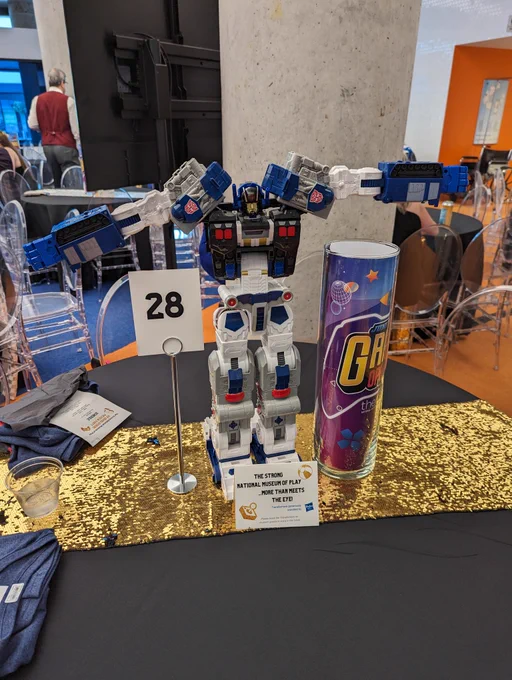 Picture of a table with a black tablecloth and a gold sash across the top. There is a Transformers Generations Legacy Series Titan Cybertron Universe Metroplex action figure arms outstretched. To the left is a sign that indicates this is table 28 and to the right is a plastic clear cylinder that has the Strong grand opening logo inside. There is a little placard that reads “The Strong National Museum of Play… More Than  Meets The Eye!”