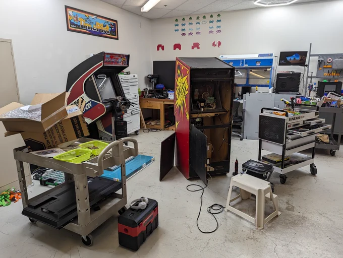 A picture of a workshop. From left to right: A cart with a monitor, parts bins, and a cardboard box on top. There is a Hang On arcade machine turned off with it’s coin door open. Above that attached to the wall is a large Time Pilot arcade marquee. There’s a white rollilng tool cart and then a bench with a drill press on top. On the wall are Space Invaders stickers. In the middle is a Stargate arcade machine facing away from us. The back is open and you can see the monitor. There’s a cordless dremel and some stepstools on the floor. In the back is a hood used in labs to extract fumes (presumably from solder). There’s a computer with monitor on a stand next to that and in front is another metallic tool cart with a few drawers open. On the far right is another cart full of parts and in the back you can see a bench with a 3D printer on top and 3D printed tool holders on the pegboard.