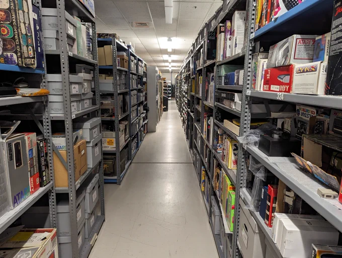 Looking down a hallway created by grey metal shelving on the right and left. They are full of boxes and other artifacts. It goes back at least 150 feet. Visible boxes in the foreground include an Odyssey, Atari 130XE, Atari Lynx, Atari 7800, various disk drives, a PS4, Guitar Hero, and then a number of pong machines. 