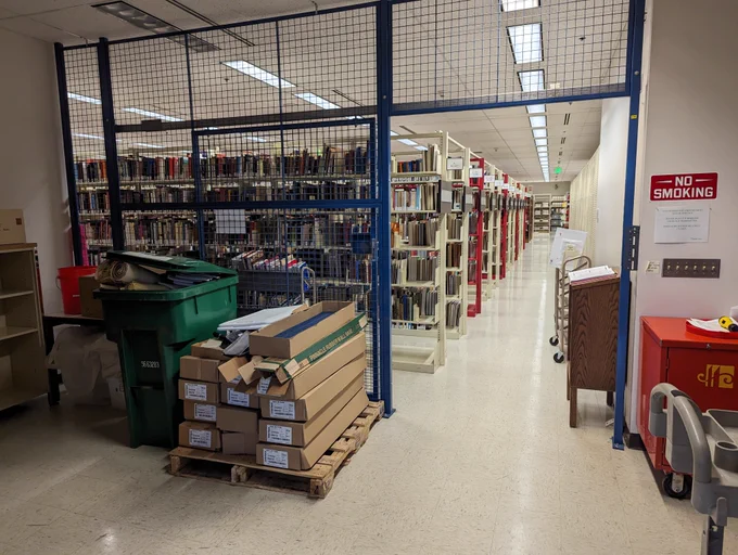 Library archive of The Strong (behind the scenes). A large blue gate is in the foreground. Visible behind it are the ends of 15-20 shelves full of printed material. There is a “No Smoking” sign prominent to the right of the gate.