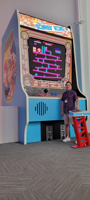 Picture of me standing in front of the large Donkey Kong arcade machine. The decorative control panel is even with my shoulders. I’m wearing a Donkey Kong shirt and shorts. In front of me is a pedestal shaped like the game’s girders with a Donkey Kong control panel on top. 
