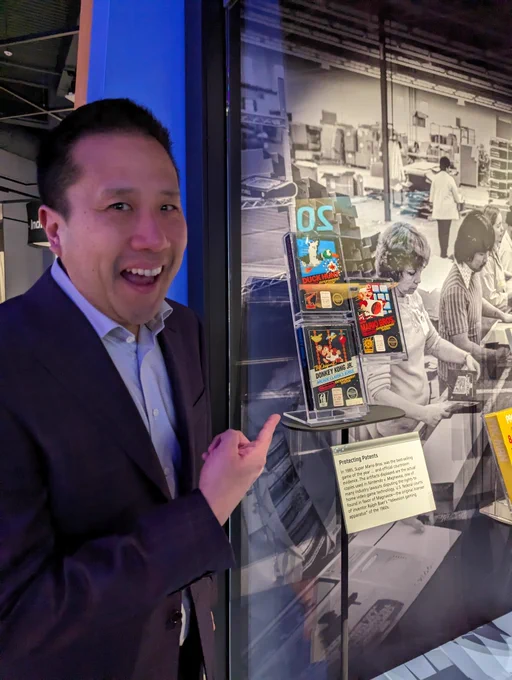 A picture of me wearing a dress shirt and sport coat excitedly pointing to my left at the three Nintendo Court Case games I donated in the museum display case. The three cartridges are Duck Hunt, Super Mario Bros, and Donkey Kong Jr. The placard underneath reads “Protecting Patents - In 1985 Super Mario Bros. was the best selling game of the year … and official courtroom evidence. The artifacts displayed are the actual copies used in Nintendo v. Magnavox, one of the many industry lawsuits disputing the rights to home video game technology. U.S. federal courts found in favor of Magnavox - the original licenser of inventor Ralph Baer’s “Television gaming apparatus” of the 1960s.