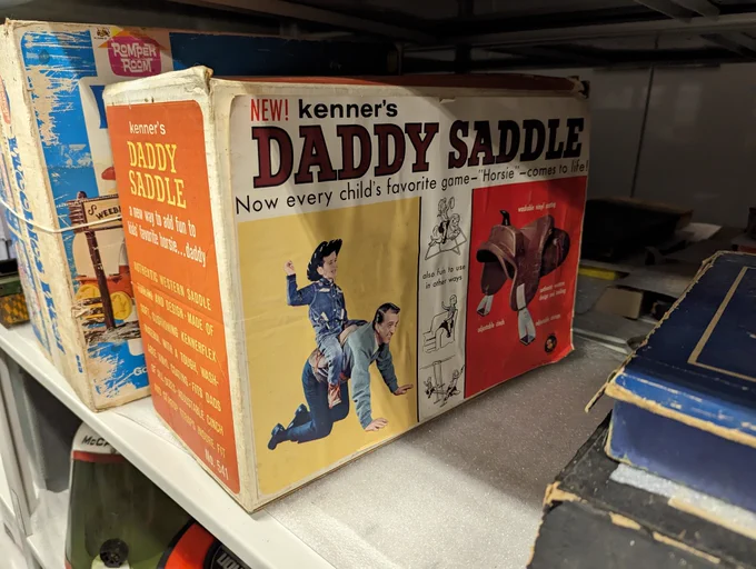A box for Kenner’s Daddy Saddle from the 1950s. The front of the box reads “New! Kenner’s Daddy Saddle - Now ever child’s favorite game - “Horsie” - comes to life!” Below that is a child in a cowboy outfit with a man underneath him on all fours wearing a small saddle. To the right is a picturee of the saddle. The middle strip reads “also fun to use in other ways” and shows the saddle being used on a rocking horse, the arm of a couch, and a seesaw. The end of the box visible reads “kenner Daddy Saddle a new way to add fun to kids’ favorite horsie… daddy” “Authentic western saddle tooling and design. Made of soft, cushioning kennerflex material with a tough, washable vinyl coating. Fits dads of all sizes. Adjustable cinch and stirrup straps to insure fit. No 541”