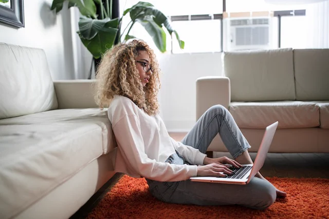 A woman sits up against a sofa with a laptop on her lap, typing. 