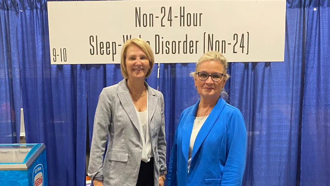 Two women stand smiling in front of a Non-24-Hour Sleep-Wake Disorder (Non-24 sign).