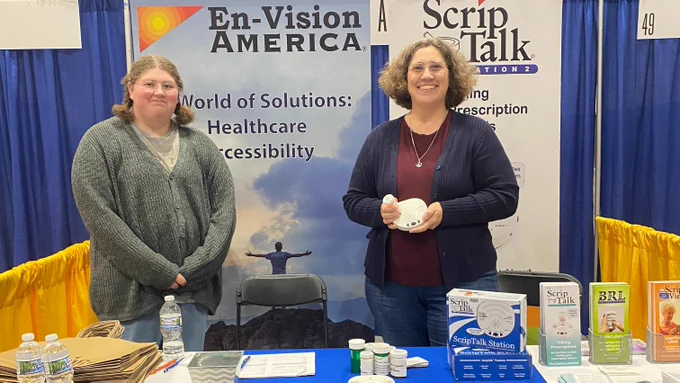 Two smiling women stand at the En-Vision America booth. One holds the Scrip Talk talking prescription label reader.
