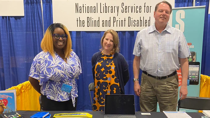 Two women and one man, all smiling, stand at the National Library Service for the Blind and Print Disabled booth.