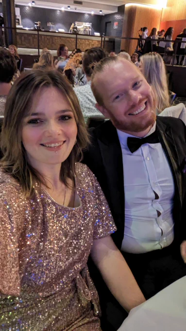 A smiling couple dressed in a gold sequined dress and a white   black tuxedo with a bowtie seated at a table.  Behind them are many other people seated for a meal.