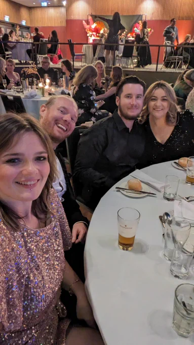 Four smiling people dressed in formalwear seated at a table with a white tablecloth.  Behind them is a room full of other tables.
