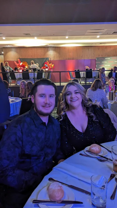 A smiling couple dressed in a floral pattern black button-up shirt and a black sequined dress seated at a table.  Behind them are other people seated at tables preparing to eat.