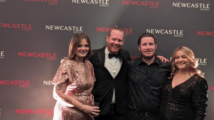 Four smiling people dressed in formal wear standing in front of a black backdrop that reads "Newcastle Weekly" in red and white text