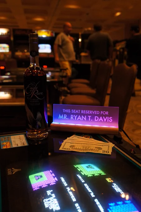 A light up sign that reads "This seat reserved for Mr. Ryan T. Davis" next to a bottle of Eagle Rare bourbon and $20 bills fanned out in front. All on top of a Warlords arcade cocktail table. In the background you can see blurry glow of other arcade marquees and monitors.