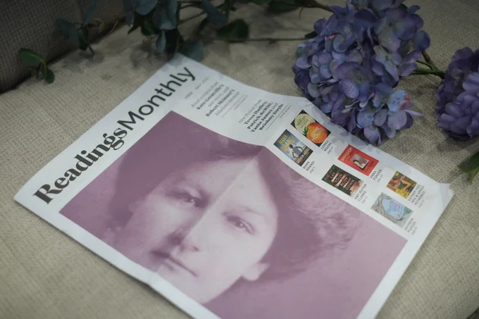 The readings monthly newsletter featuring a detail from the book Restless Dolly Maunder: a black and white photo depicting a 19th century woman's face. The newsletter is displayed with an assortment of purple flowers and eucalypt leaves