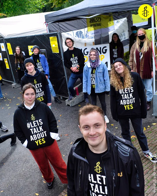 A group of Amnesty activists standing outside in the rain next to an Amnesty stand look at the camera. All are wearing jumpers saying '#Let's talk about yes' with Amnesty logos on