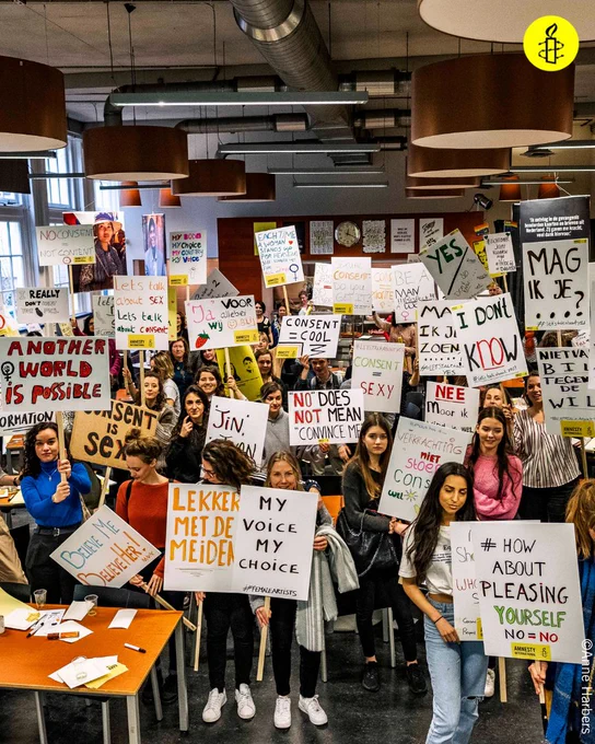 Tens of activists hold placards with slogans in Dutch and English about consent on them face the camera. One in Dutch reads 'Mag ik je...?'. One in English reads 'How about pleasing yourself? No = No'