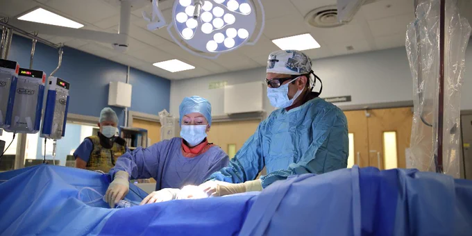 Dr Nayak is wearing blue scrubs and assited by another person doing a thrombectomy. The table is covered in blue drapes and a large surgical light is overhead. 