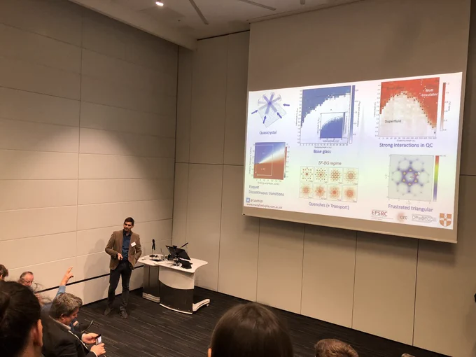 Photo of Ulrich standing in front of a podium at the front of a lecture theatre with a large projector screen covered with various experiment images behind him. He’s currently listening to an audience question.
The lecture theatre carpet is dark grey and the walls are white panels - it’s much nicer in real life than it looks in the photo!