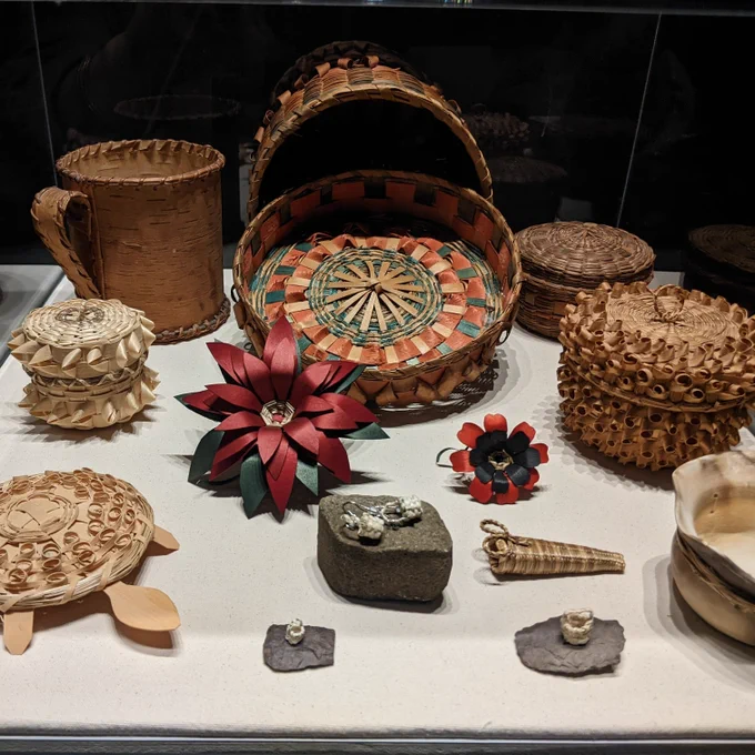 Photograph of Mi'kmaw baskets and wooden flowers in display case. Some items include a wooden poinsettias, tiny baskets the size of your pinky finger, and a turtle shaped basket. 