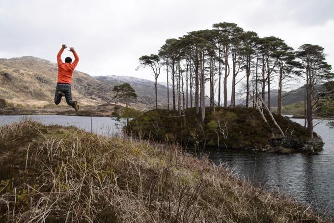 A man jumping high in the air off a hillock above a loch. There is an island in the middle of the loch covered in thin trees. The hillock in the foreground and the hills in the background are treeless, with rough grass seen on the hillock where the man is jumping. The man is wearing a bright orange top.