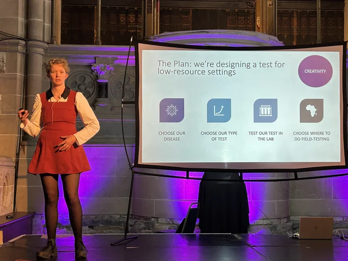 PhD student Ella presenting on stage in Cottiers with her PowerPoint slides displayed behind her.