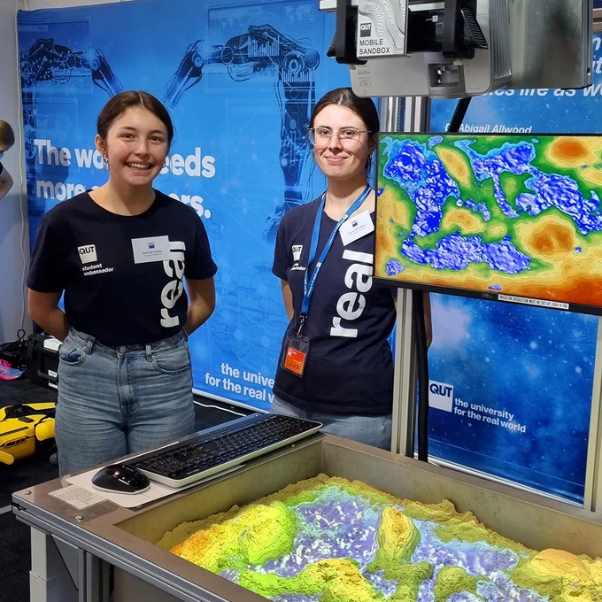 2 x young females standing next to engineering showcase wearing QUT real world tshirts and smiling at the camera.