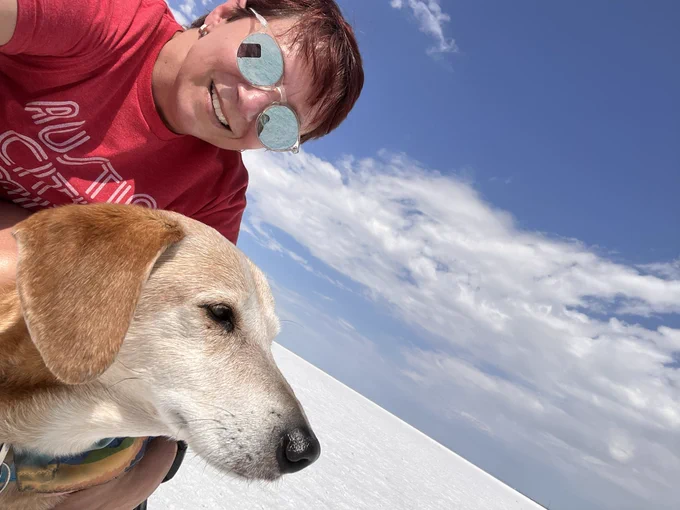 Woman in sunglasses and red Austin City Limits tshirt, and brown dog with grey face on the white salt flats with blue sky and white clouds above
