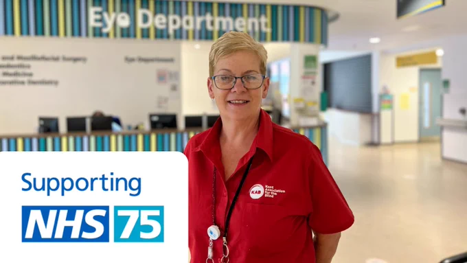 Sue stands in the reception area of the eye department within the hospital. Supporting NHS 75 logo appears bottom left.