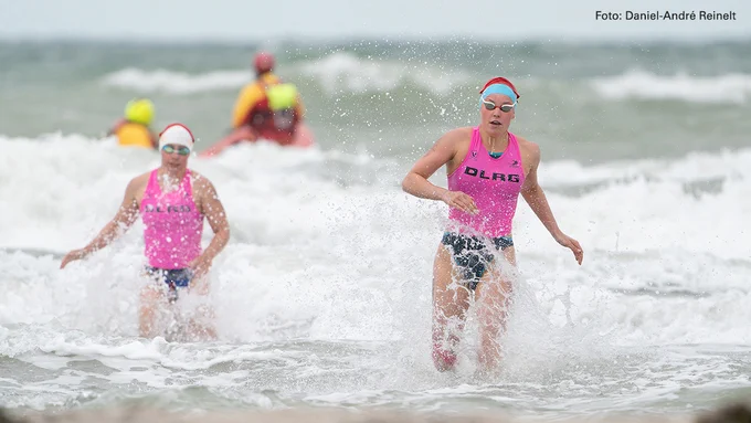 Zwei Rettungssportlerinnen der DLRG laufen durch die Brandung in Richtung Strand.