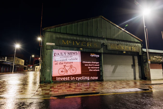 Projection on the side of a warehouse in Glasgow. Image shows newspaper clipping with a picture of a bike and headline: "November, 1839. Revealed: vehicle of the future." Additional text reads "Climate change: we don't need to reinvent the wheel. Invest in cycling now."