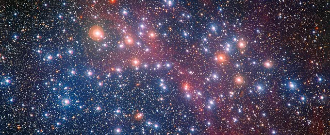 An image of the star cluster NGC 3532. Stars of blue, orange and white shine brightly against a dark red and black backdrop of space. Credit: ESO