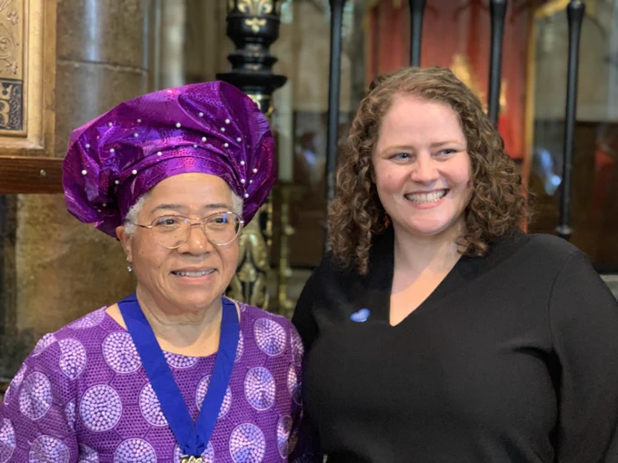 Dame Elizabeth Anionwu, in a purple dress with purple head wrap, next to Eloise. Both are smiling and looking off camera.
