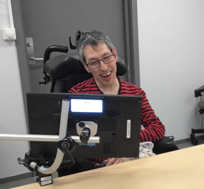 This is an image of Ahmad Naziri. In the photo, Ahmad is wearing a black and red striped long sleeve t-shirt and glasses. Ahmad is pictured in a wheelchair seated at a table with an AAC device screen in front of him.