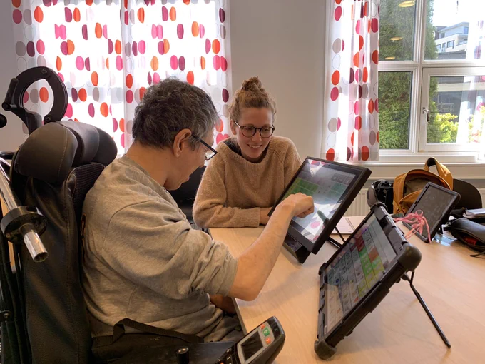 This is an image taken from the side of Ahmad Naziri using two AAC devices on the table in front of him. Next to him is a woman with glasses assisting him with using the AAC devices.