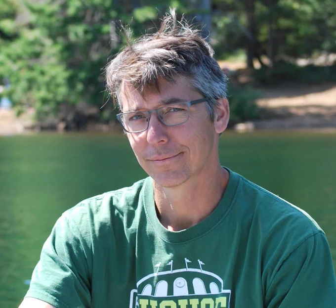 This is an image of Chris Gibbons, the author of the piece. Chris is a white man with glasses wearing a green t-shirt. In the photo, he is pictured outdoors in front of a body of water with trees and dirt visible on the opposite side of the water behind.