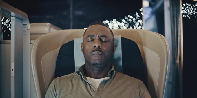 Idris Elba as Sam Nelson. He is wearing an olive green quarter-button long sleeve sweater with a white t-shirt underneath. He is sitting in a first class airplane seat with his eyes closed. The tone is serene.