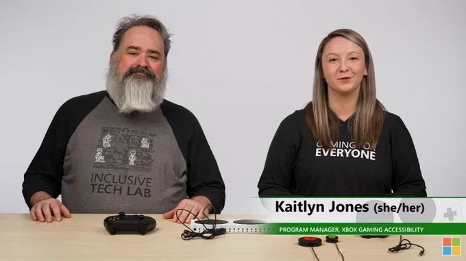 two people stand behind a desk on which on which there is an Xbox adaptive controller and peripheral The first is a middle aged white man with a gray bushy beard wearing a inclusive tech lab teacher the second is a young woman with blonde hair wearing a gaming for everyone hoodie