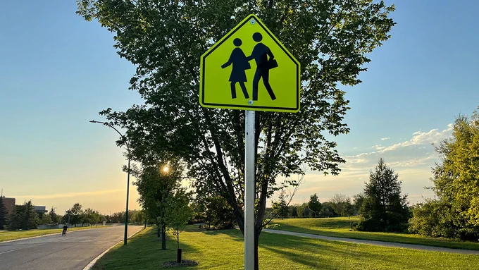 Yellow school zone sign without a posted speed limit along a park and grassy area with trees next to a road. 