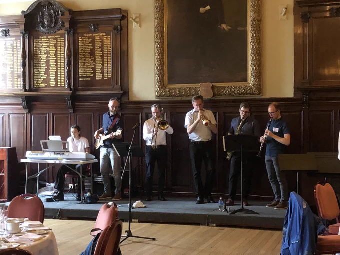 A band of six physicists are standing playing music on a small stage at the front of a beautiful wood panelled room set for dinner.