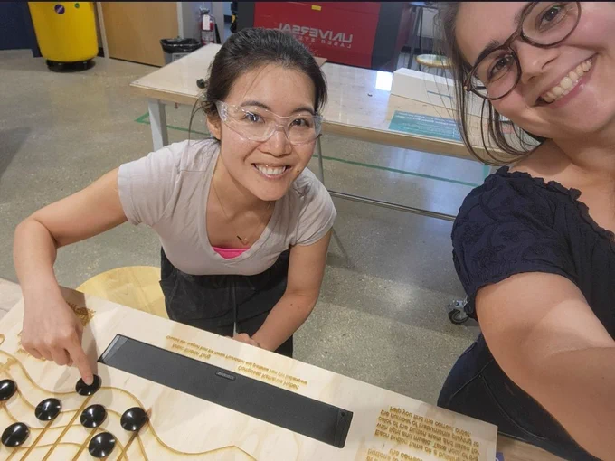 Sharon (left) and Kat(right) smiling after their victories. Sharon designed a blueprint for and cut out the control panel, while Kat designed the art/text on top of the control panel and used a laser cutter to raster the design into the wood. Sharon is pressing a black button in the photo. 