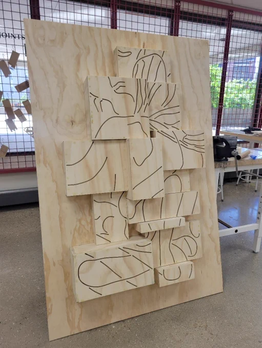 Large wooden plywood panel with 13 3D smaller boxes that protrude from the large panel at various depths. The 13 boxes have designs cut into them. 