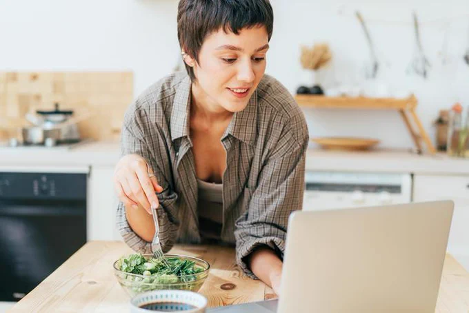 A woman sits at a desk, plunging a fork into a bowl of salad with one hand, typing at a laptop with another, a smile playing across her face