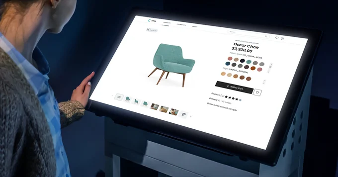 Photo of a woman looking at a big screen. The screen is showing the product page of an "Oscar Chair", with the image of the chair to the left and the product configuration (color, material) to the right.