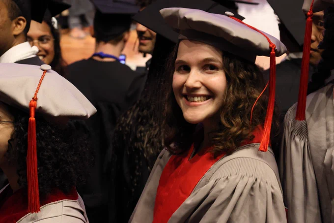 Person in gray and maroon PhD graduation robes with a graduation hat at a graduation ceremony.