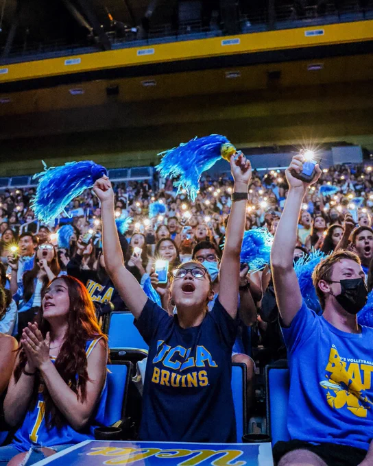 A fan cheers with pom poms, and behind in the stands are rows of fans holding up cell phone lights during UCLA’s pregame introductions.